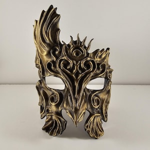 May include: A detailed, gold-colored masquerade mask with an ornate design. The mask features eye openings, intricate scrollwork, and a stylized crown-like element. The overall aesthetic is reminiscent of fantasy or historical themes.