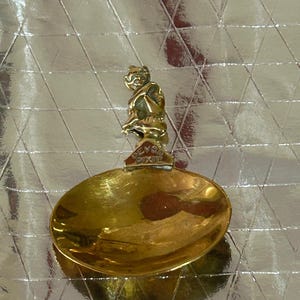 May include: A gold-coloured spoon featuring a pixie figure on the handle. The spoon has an oval bowl and a decorative handle with a small pixie figure. The handle is engraved with the words "Devon Pixie".