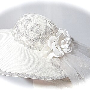 Puede incluir: Un sombrero de sol blanco de ala ancha, adornado con un delicado encaje y una gran flor blanca. El sombrero tambi&eacute;n tiene un velo blanco transparente.