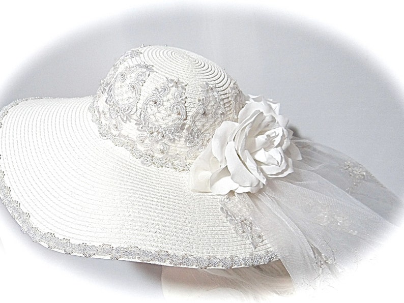 Puede incluir: Un sombrero de sol blanco de ala ancha, adornado con un delicado encaje y una gran flor blanca. El sombrero tambi&eacute;n tiene un velo blanco transparente.