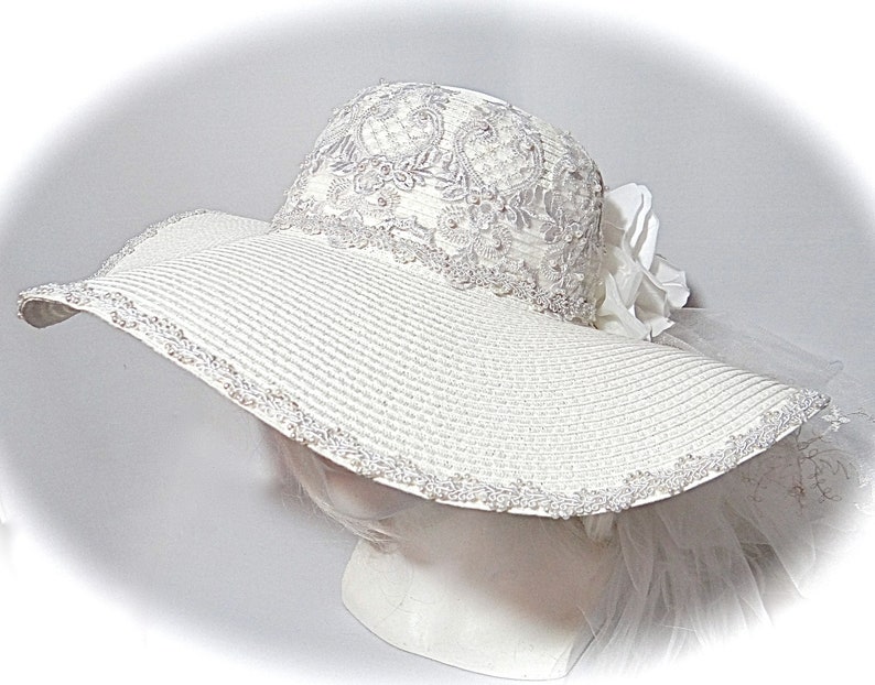 Puede incluir: Un sombrero blanco de ala ancha para el sol con un borde de encaje y un adorno de flor blanca.