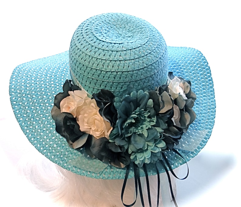 May include: A light blue straw sun hat with a wide brim and a large flower embellishment. The flower is made of white, black, and teal fabric.