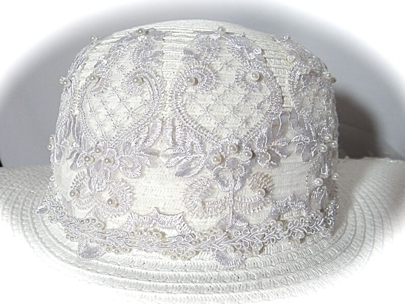 Puede incluir: Sombrero de sol blanco con ala ancha y adorno de encaje blanco con detalles de perlas.