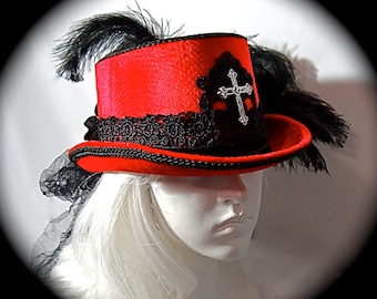 May include: A red top hat with a black lace trim and a silver cross embellishment. The hat is adorned with black feathers.