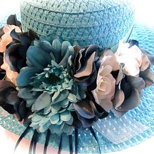 May include: A light blue straw hat with a wide brim and a large flower embellishment. The flower is made of white, blue, and dark blue fabric.