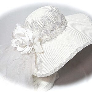 Puede incluir: Sombrero blanco de ala ancha con una flor blanca y un velo blanco. El sombrero est&aacute; decorado con encaje blanco y cuentas.