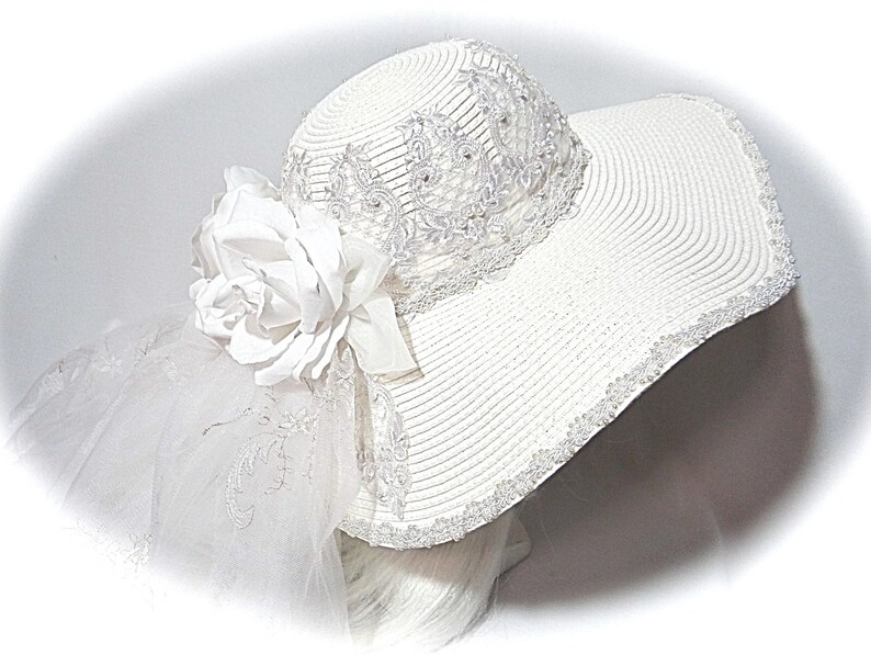 Puede incluir: Sombrero blanco de ala ancha con una flor blanca y un velo blanco. El sombrero est&aacute; decorado con encaje blanco y cuentas.