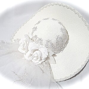 Puede incluir: Un sombrero de sol blanco con ala ancha y un ribete de encaje blanco. El sombrero tiene un adorno de flores blancas en un lado.
