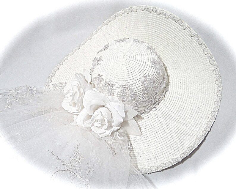 Puede incluir: Un sombrero de sol blanco con ala ancha y un ribete de encaje blanco. El sombrero tiene un adorno de flores blancas en un lado.