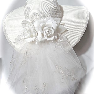 Puede incluir: Sombrero blanco de ala ancha con velo blanco y adorno de flores blancas.
