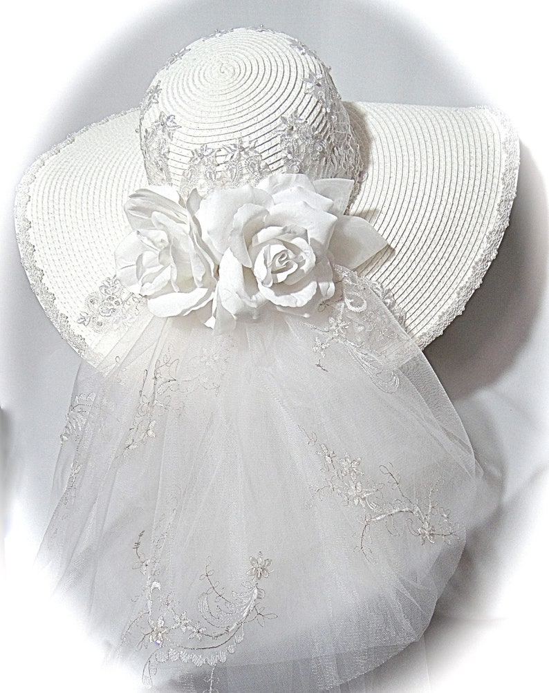 Puede incluir: Sombrero blanco de ala ancha con velo blanco y adorno de flores blancas.