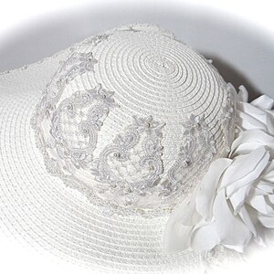 Puede incluir: Sombrero de sol blanco con ala ancha y ribete de encaje blanco. El sombrero est&aacute; decorado con una flor blanca.