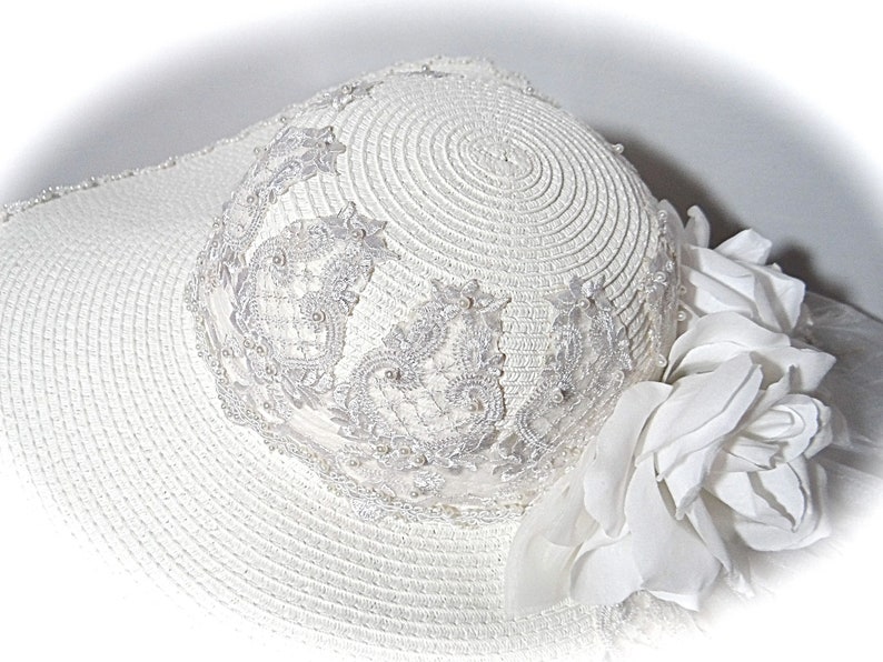 Puede incluir: Sombrero de sol blanco con ala ancha y ribete de encaje blanco. El sombrero est&aacute; decorado con una flor blanca.