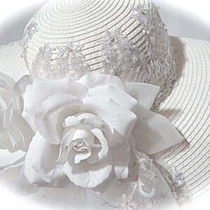 Puede incluir: Un sombrero de paja blanco con ala ancha y ribete de encaje blanco. El sombrero est&aacute; decorado con una gran flor blanca.