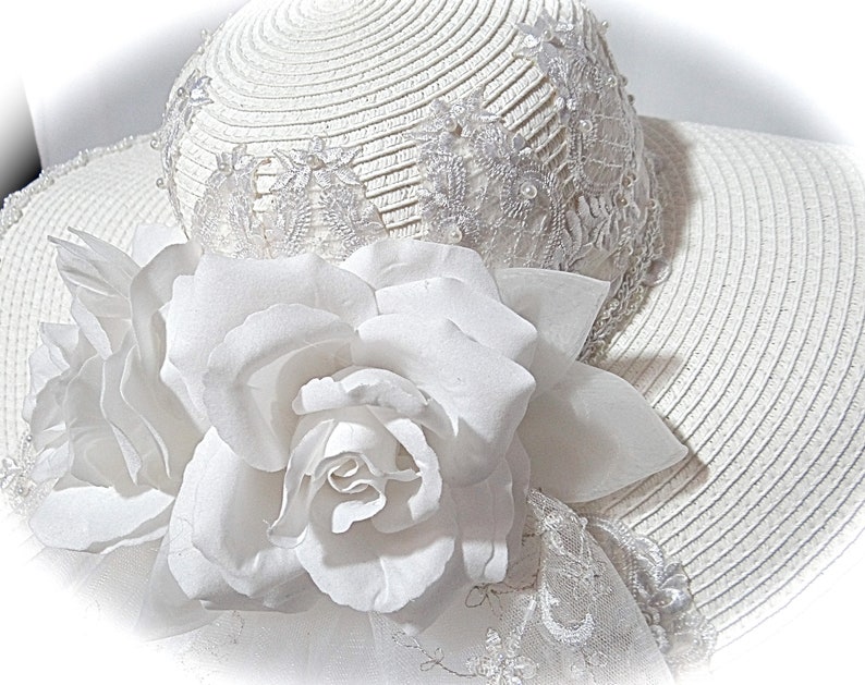 Puede incluir: Un sombrero de paja blanco con ala ancha y ribete de encaje blanco. El sombrero est&aacute; decorado con una gran flor blanca.