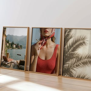 May include: Three framed art prints leaning against a white wall. The prints feature a scenic lake view, a woman in a red swimsuit and headscarf, and a palm leaf shadow. The frames are a light wood color.