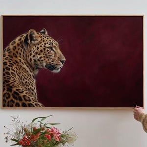 May include: A rectangular wall art print featuring a profile view of a leopard with golden fur and black spots. The leopard is positioned on the left side against a solid, deep burgundy background. The artwork is encased in a thin wooden frame.