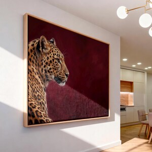 May include: A rectangular wall art piece featuring a profile portrait of a leopard with golden fur and black spots. The leopard is positioned on the left side against a deep, solid burgundy background. The artwork is encased in a light wood frame.