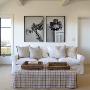 May include: A living room scene featuring a white sofa with beige pillows and a plaid ottoman. Two framed black and white art prints hang above the sofa. Woven trays sit on the ottoman, and a window is visible on the left.