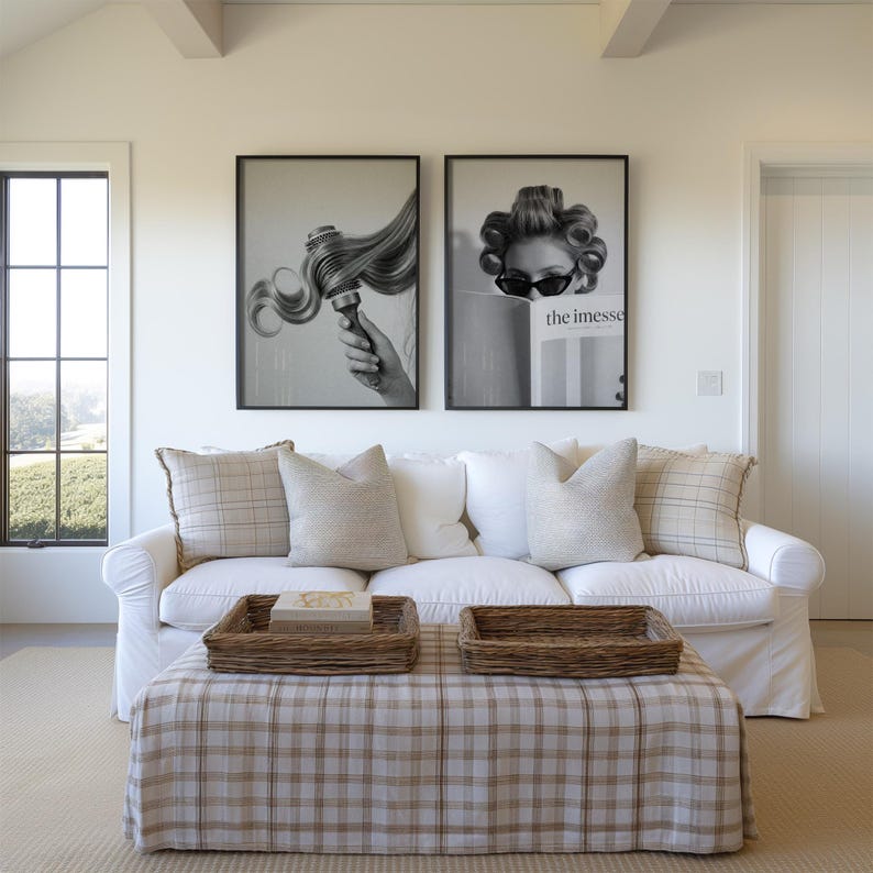 May include: A living room scene featuring a white sofa with beige pillows and a plaid ottoman. Two framed black and white art prints hang above the sofa. Woven trays sit on the ottoman, and a window is visible on the left.