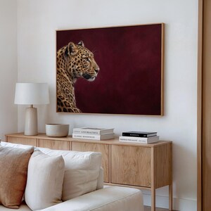 May include: A rectangular wall art print featuring a leopard in profile against a deep burgundy background, displayed in a thin wooden frame above a light oak sideboard with books, a ceramic bowl, and a white table lamp.