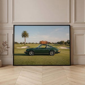 May include: Framed print of a dark green vintage sports car on a golf course, with a suitcase on the roof. The image is set against a blue sky, with a palm tree and golf course features in the background.