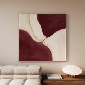 May include: A square wall art piece featuring large, fluid abstract shapes in deep burgundy and off-white. A thin, delicate gold line curves through the center, separating the two primary color blocks. The composition is balanced and symmetrical.