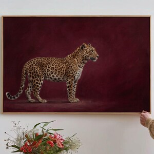 May include: A rectangular wall art print featuring a realistic leopard standing in profile against a solid, deep burgundy background. The leopard has a golden-tan coat covered in black rosettes and spots. The artwork is enclosed in a thin wooden frame.