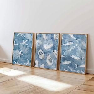 Puede incluir: Tres impresiones artísticas enmarcadas en acuarela con vida marina. Las impresiones muestran estrellas de mar, ostras y peces sobre un fondo azul. Cada impresión está en un marco de madera, creando un tema costero cohesivo.