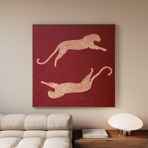 May include: A square wall art piece featuring a deep red background with two leaping leopards depicted in a metallic gold finish. The leopards are positioned in a mirrored orientation, one above the other, showcasing detailed spotted patterns.