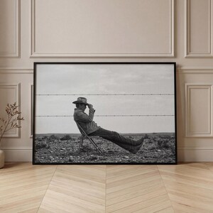 May include: Black and white framed print of a person sitting in a chair outdoors. The person is wearing a hat and jacket, resting against a barbed wire fence. The background shows a cloudy sky and a barren landscape.