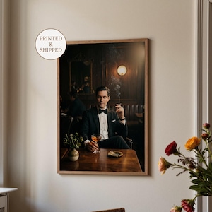 Pode incluir: Uma impressão emoldurada retrata uma pessoa de smoking preto, sentada em uma mesa de madeira em um bar com pouca luz. A pessoa segura um coquetel e um cigarro aceso. Um pequeno vaso e um cinzeiro estão sobre a mesa. A impressão está rotulada "PRINTED & SHIPPED".