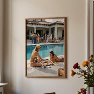 Könnte beinhalten: Gerahmtes Foto von zwei Personen, die am Pool Backgammon spielen. Eine Person trägt einen gelben Bikini und ein Kopftuch, die andere einen gemusterten Bikini. Im Hintergrund ist eine Gruppe von Personen zu sehen. Das Foto ist in einem Holzrahmen.