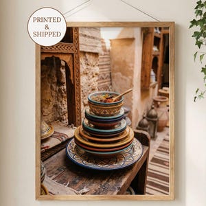May include: A framed print featuring a stack of colorful ceramic bowls and plates with intricate patterns. A wooden spoon rests in the top bowl. The print is displayed in a wooden frame and includes the text "PRINTED & SHIPPED".