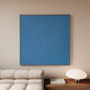 May include: A square wall art piece featuring a solid blue background with small, scattered figures in black, red, and white. The painting is framed in a thin, light-colored wood frame and is displayed on a wall above a piece of furniture.