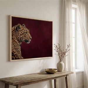 May include: A rectangular wall art print featuring a profile view of a leopard with golden fur and black spots, set against a deep burgundy background. The artwork is enclosed in a light-colored wooden frame, showcasing the animal's head and neck.