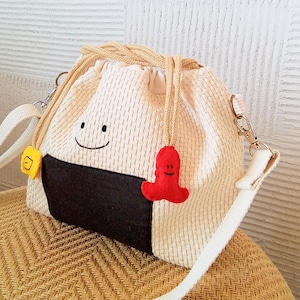 以下が含まれることがあります： おにぎり型のかわいいベージュの織りショルダーバッグ。黒い海苔のアクセントと笑顔の顔が特徴です。ベージュの巾着開閉、白いストラップ、赤いチャームが付いています。バッグには黄色の正方形のタグが付いています。