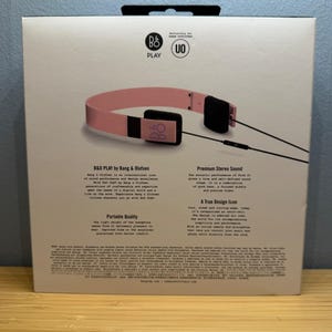 May include: A box featuring pink B&O PLAY headphones. The headphones have a pink headband and black earcups. The box has text that reads "B&O PLAY by Bang & Olufsen" and "Premium Stereo Sound".
