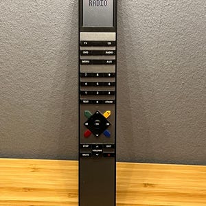 Può includere: Un telecomando nero Bang & Olufsen con uno schermo che mostra "RADIO". Il telecomando ha pulsanti per TV, CD, DVD, Radio, Menu, Aux, i numeri 0-9 e altre funzioni. È appoggiato su una superficie di legno.