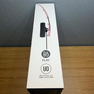 May include: A white box featuring a pink and black headset design. The box has the text "B&O PLAY" and "UO" with the words "Exclusively for URBAN OUTFITTERS". The box is on a wooden surface.
