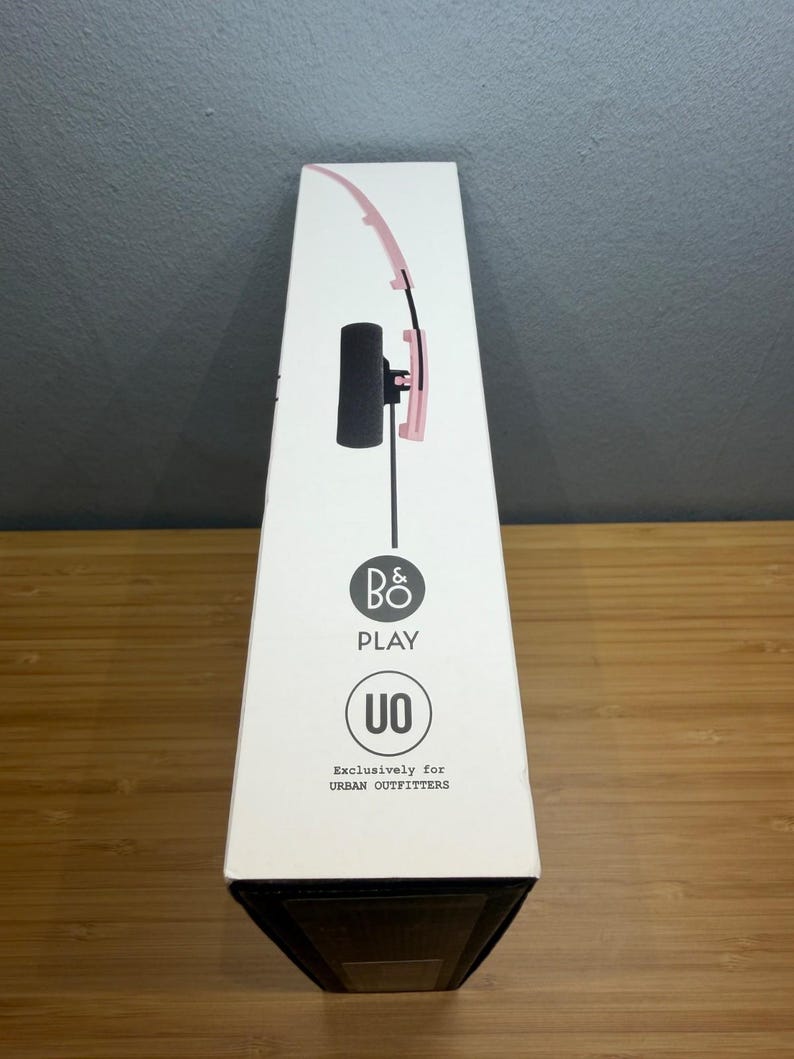 May include: A white box featuring a pink and black headset design. The box has the text "B&O PLAY" and "UO" with the words "Exclusively for URBAN OUTFITTERS". The box is on a wooden surface.