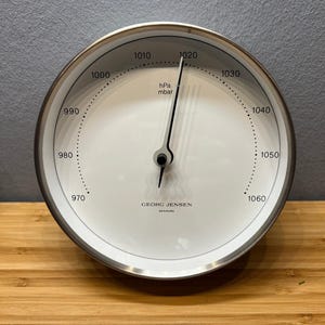 May include: A round, silver-rimmed barometer with a white face. The face displays numbers from 970 to 1060, with a black needle indicating the current pressure. The text "GEORG JENSEN DENMARK" is printed at the bottom.