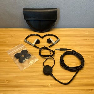 Può includere: Auricolari neri con dettagli argentati, una custodia in pelle nera e auricolari di ricambio. Le cuffie hanno un cavo nero e un jack audio da 3,5 mm. La custodia è semicircolare e ha il nome del marchio stampato sopra.