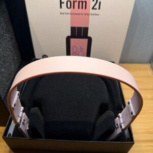 May include: A pair of pink headphones with a black interior, presented in a black box. The box features the text "Form 2i" and "B&O PLAY Exclusively for Urban Outfitters" in black and pink.
