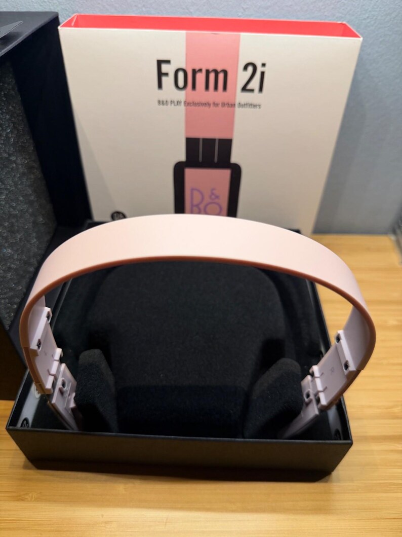 May include: A pair of pink headphones with a black interior, presented in a black box. The box features the text "Form 2i" and "B&O PLAY Exclusively for Urban Outfitters" in black and pink.