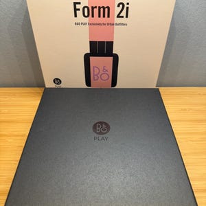 May include: A black box with the logo "B&O PLAY" and the text "Form 2i" on the front. The box is sitting on a wooden surface. The top box has a pink stripe and the text "B&O PLAY Exclusively for Urban Outfitters".
