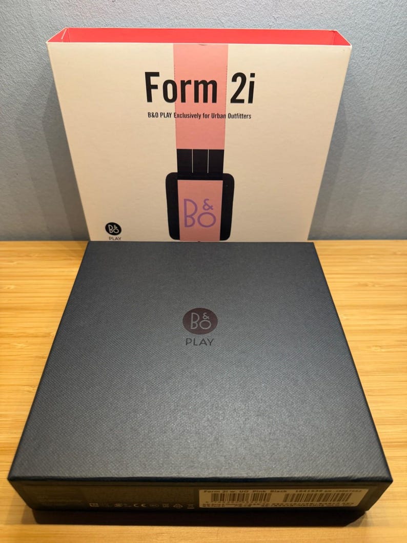 May include: A black box with the logo "B&O PLAY" and the text "Form 2i" on the front. The box is sitting on a wooden surface. The top box has a pink stripe and the text "B&O PLAY Exclusively for Urban Outfitters".