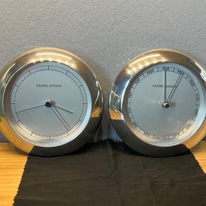 Könnte beinhalten: Zwei silberfarbene Georg Jensen Uhren mit hellgrauen Zifferblättern und schwarzen Zeigern. Die Uhren haben ein kreisförmiges Design mit einer silbernen Lünette. Eine Uhr zeigt die Uhrzeit, die andere den Luftdruck an.