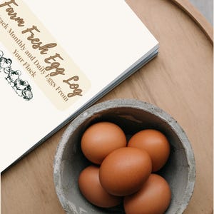 May include: A concrete bowl filled with five brown eggs sits on a wooden surface next to a notebook. The notebook has a label that reads "Farm Fresh Egg Log" with text below it.