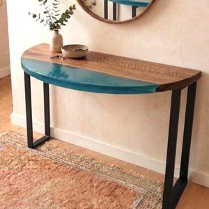 May include: A half-moon console table with a wooden top and teal resin inlay. The table has black metal legs and is placed against a light-colored wall. A round mirror hangs above the table, reflecting the table's surface. The text "Craft Studio by Arhan" is visible on the table.